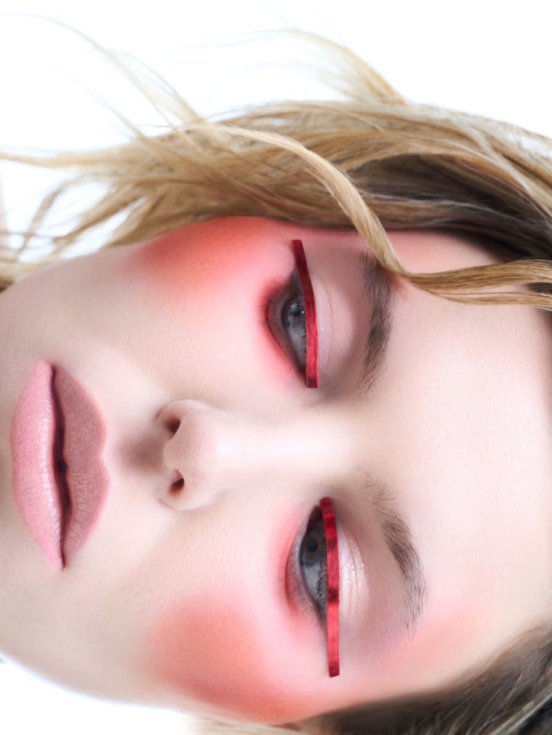 Close-up beauty portrait of a young woman with artistic red makeup, including blush and graphic lines around her eyes, on a white background.