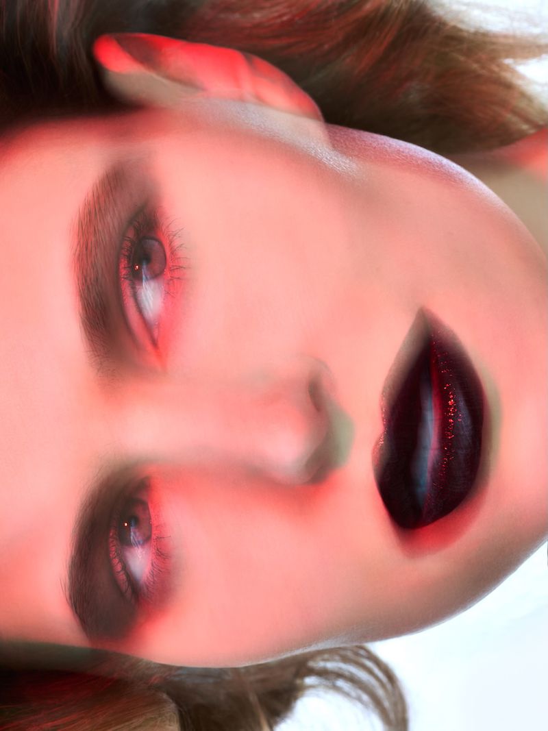 Close-up portrait of an adult female with red-toned soft lighting, dark lipstick, and soft focus against a white background.