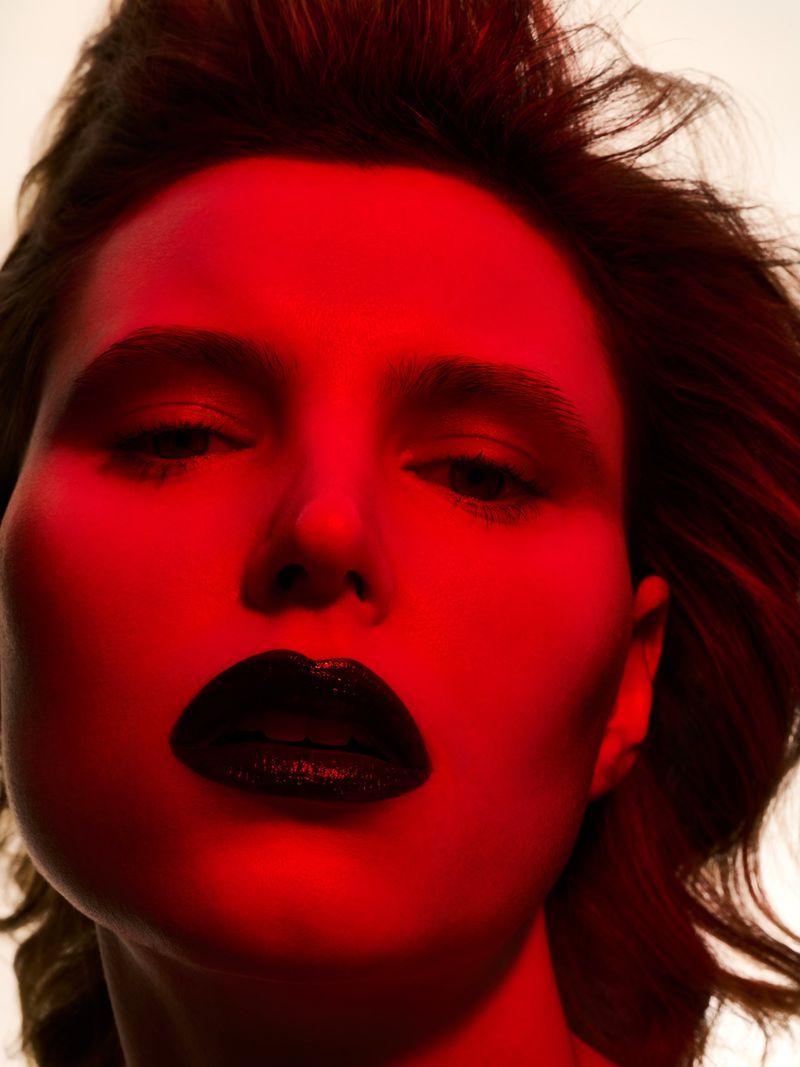 Close-up portrait of a woman with dark lipstick, illuminated by red lighting, against a light background, for beauty and fashion purposes.