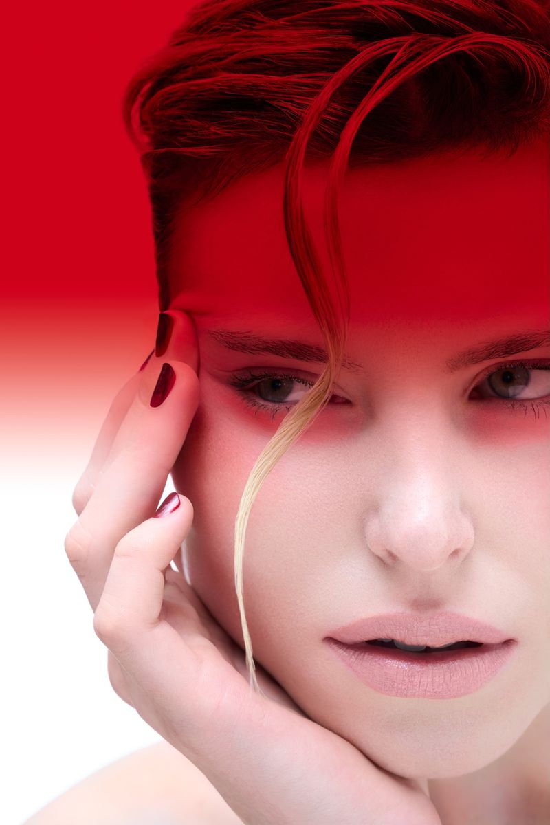 Close-up portrait of a young adult woman with red-tinted hair, her hand resting on her face, with red and white background.