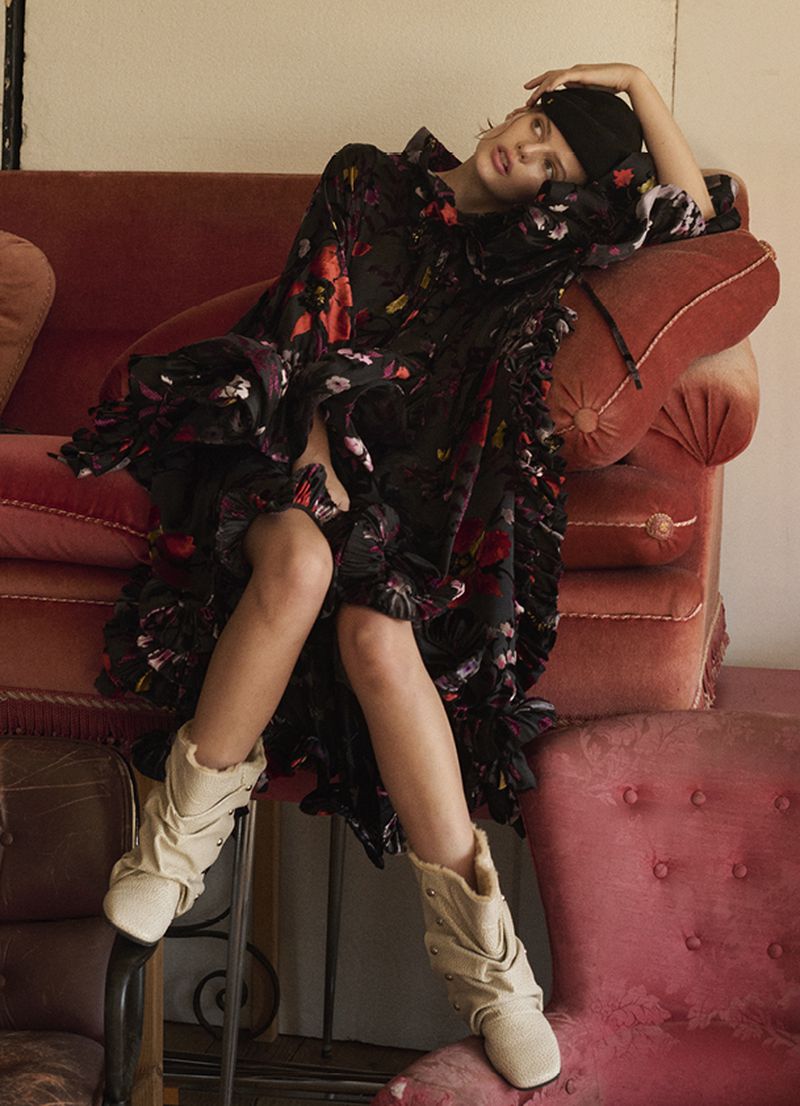 A woman in a black floral dress and beige boots poses on vintage furniture, against a neutral backdrop for a fashion editorial.