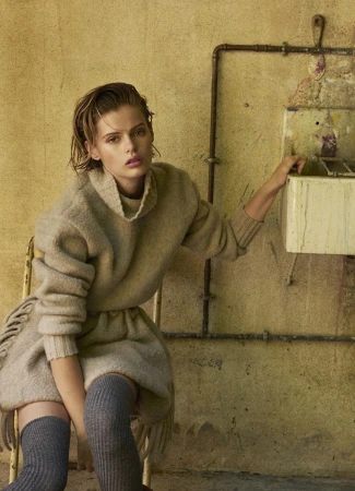 A young woman with wet hair and a turtleneck sweater dress sitting near an old sink with peeling paint in a rustic indoor setting.