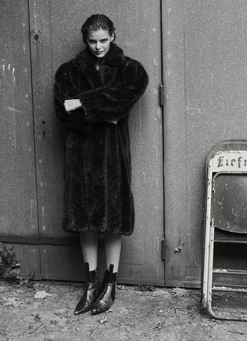 Full-length portrait of an adult woman in a dark fur coat and black boots standing against a textured metal wall, black and white photo.