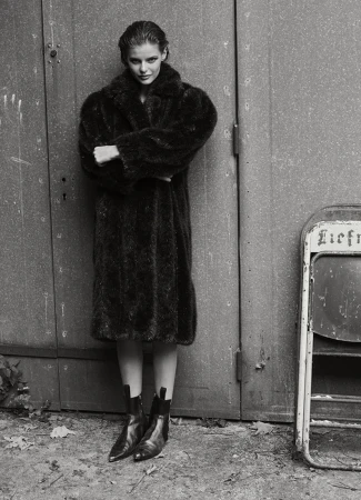 Full-length portrait of an adult woman in a dark fur coat and black boots standing against a textured metal wall, black and white photo.