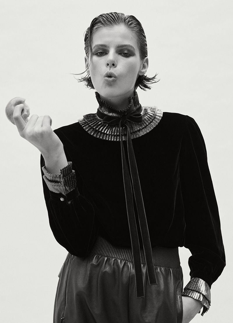Studio shot of a young adult woman with dark hair, wearing a black velvet top and pants, holding an apple against a white backdrop.
