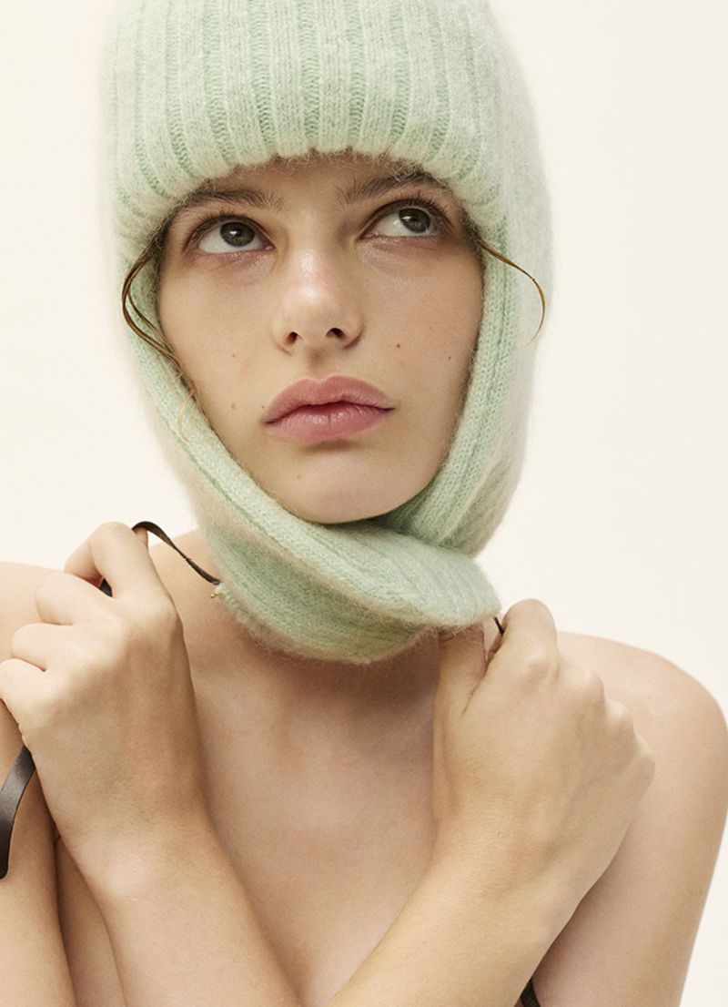 Close-up studio portrait of a light-skinned young adult woman wearing a mint green wool balaclava with a neutral facial expression looking upward.