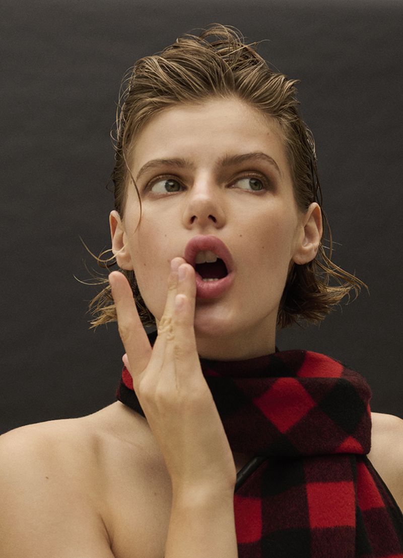 Close-up portrait of a model with wet hair, open mouth and hand near face, wearing a red and black buffalo plaid scarf on a dark background.