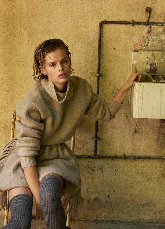 Aria Shine in a beige fringed knit sweater dress and grey thigh-high socks seated beside an industrial wall sink.