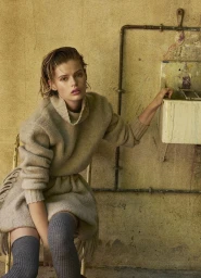 Aria Shine in a beige fringed knit sweater dress and grey thigh-high socks seated beside an industrial wall sink.