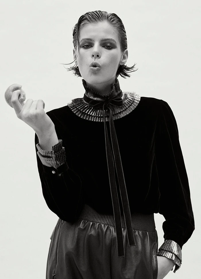 Model with slicked-back dark hair wearing a ruffled velvet blouse against a white background