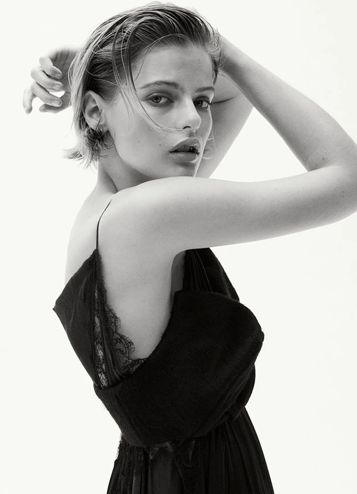 Black and white portrait of a woman looking over her shoulder wearing a black lace-trimmed strap dress against a white background.