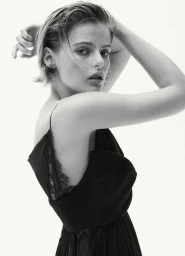 Black and white portrait of a woman looking over her shoulder wearing a black lace-trimmed strap dress against a white background.