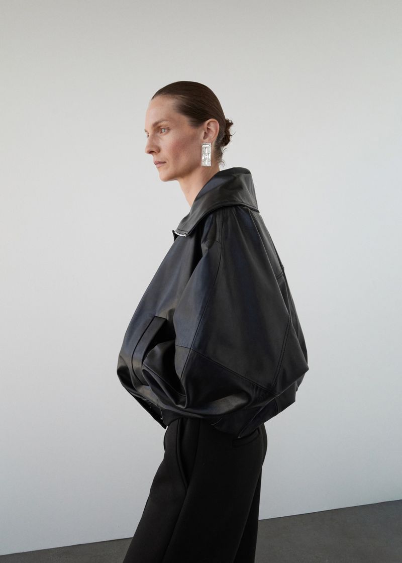 Studio shot of a woman in a black leather jacket and black pants posing in profile against a plain white background.