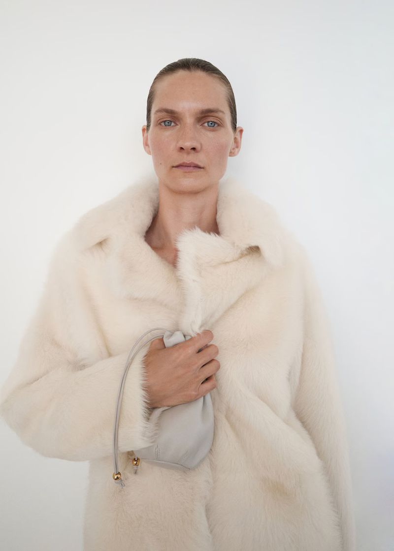Headshot of a fair-skinned adult female model wearing a cream-colored fur coat and holding a light grey pouch against a white backdrop.