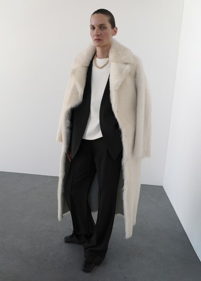 A full-length shot captures an adult female in a white faux fur coat over a black suit and T-shirt, standing in a bright white studio.