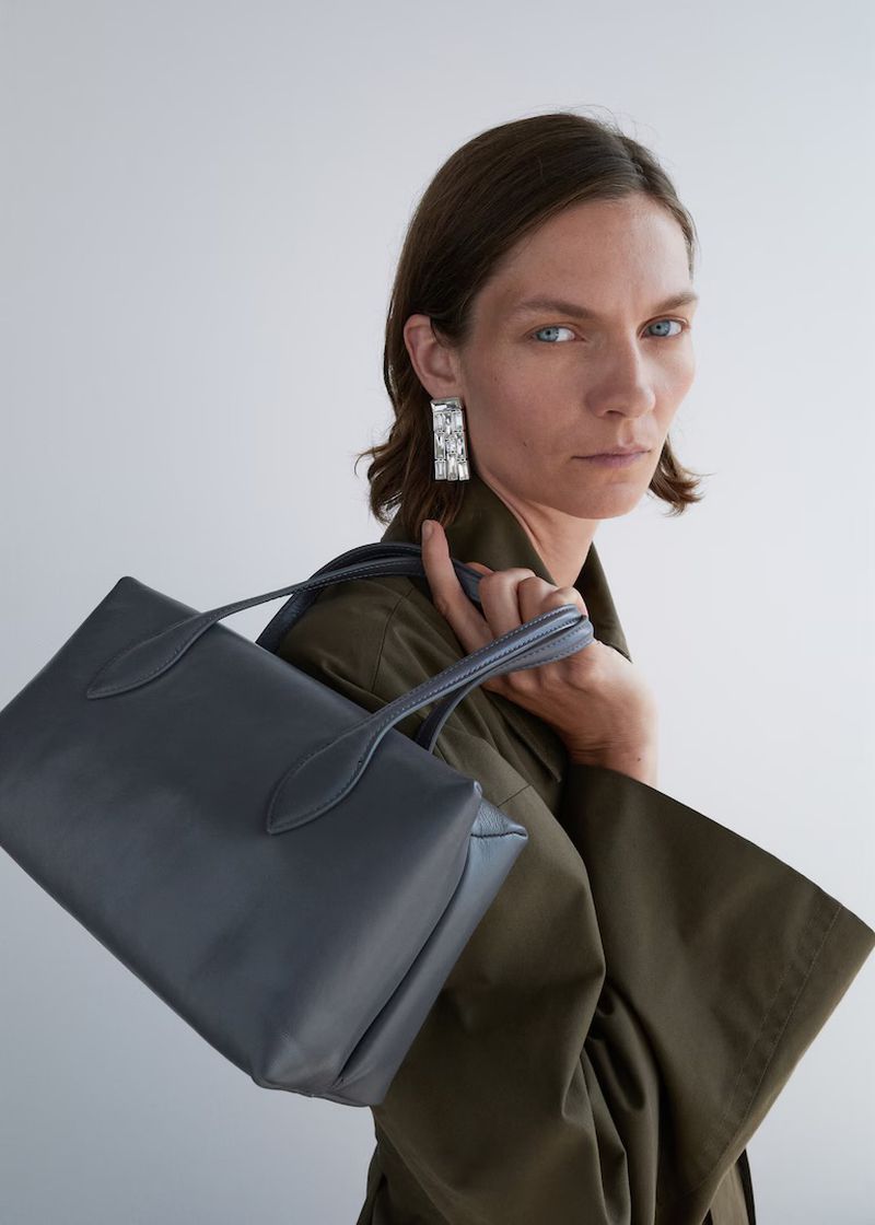 Model looking over her shoulder while carrying a structured slate grey leather handbag and wearing an olive green wide-sleeve coat.