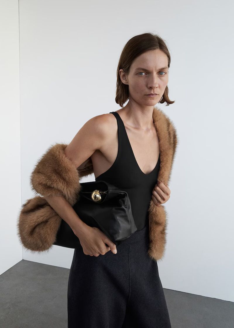 Woman in black bodysuit holding a leather handbag with a fur stole, posing in a bright studio setting. Fashion photography.