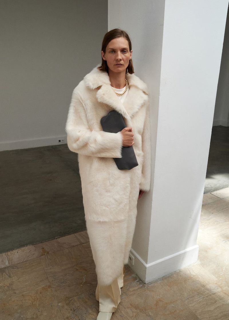 A woman in an off-white coat and pants poses indoors, holding a grey clutch bag, with a pillar behind her in a fashion-style image.