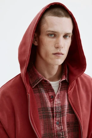 Model Jake Hodder wearing a red hooded sweatshirt over a red plaid flannel shirt against a plain white studio background.