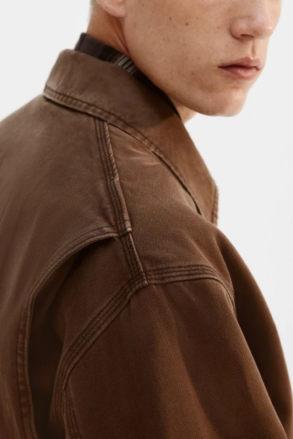 Cropped view of a fair-skinned man wearing a brown collared jacket with prominent seams against a clean white background.