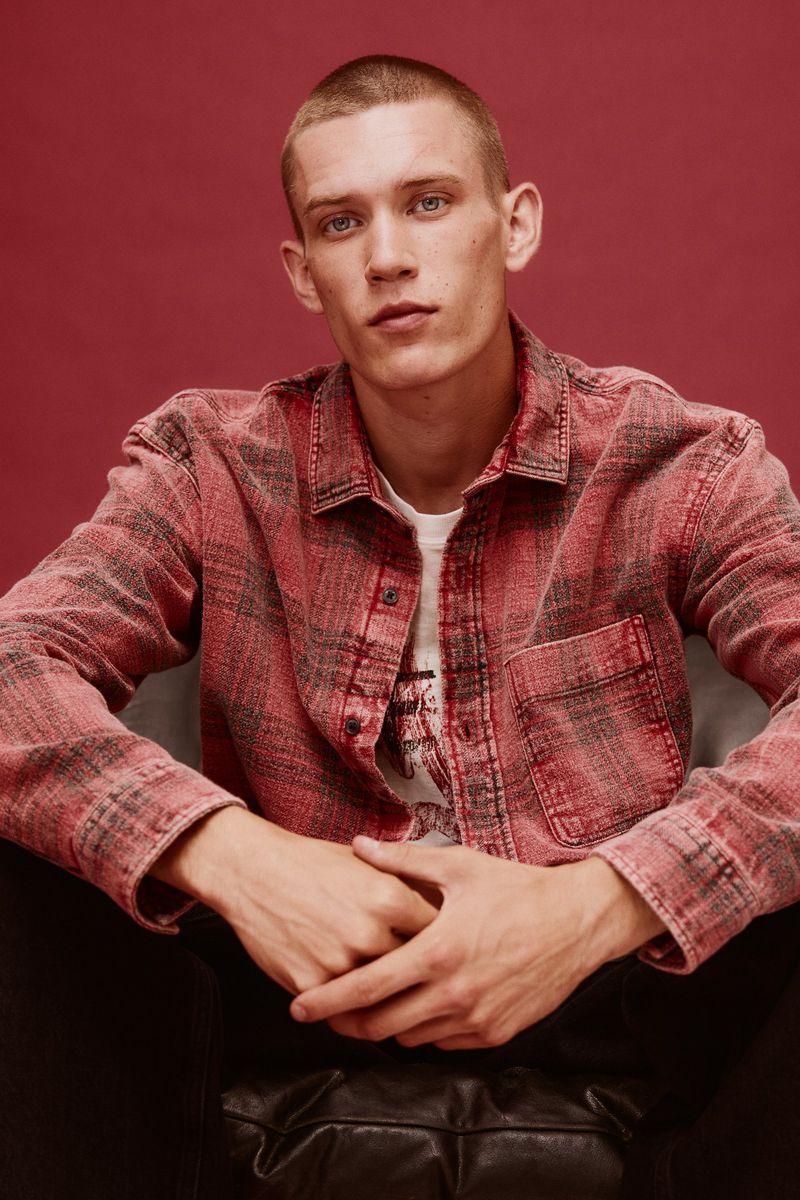 Young adult man with short hair poses in a pink and black flannel shirt, sitting in a chair against a dark pink background.