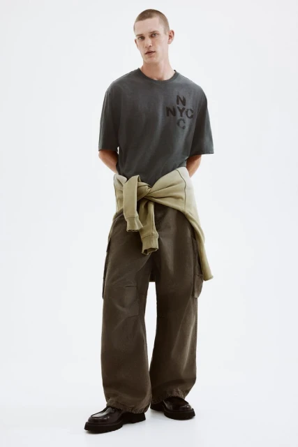 A young adult male stands in a full shot, wearing a grey t-shirt with 'NYC' printed on it, olive green pants, and black shoes, against a white background.