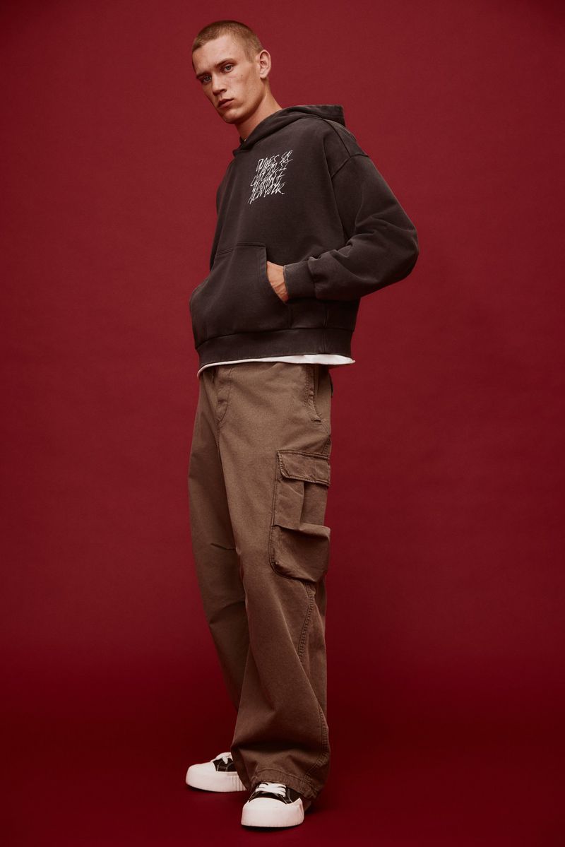A young adult male is posing indoors wearing a grey hoodie, brown cargo pants, and white sneakers against a dark red background.