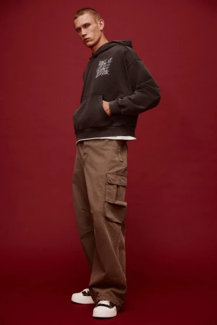 A young adult male is posing indoors wearing a grey hoodie, brown cargo pants, and white sneakers against a dark red background.