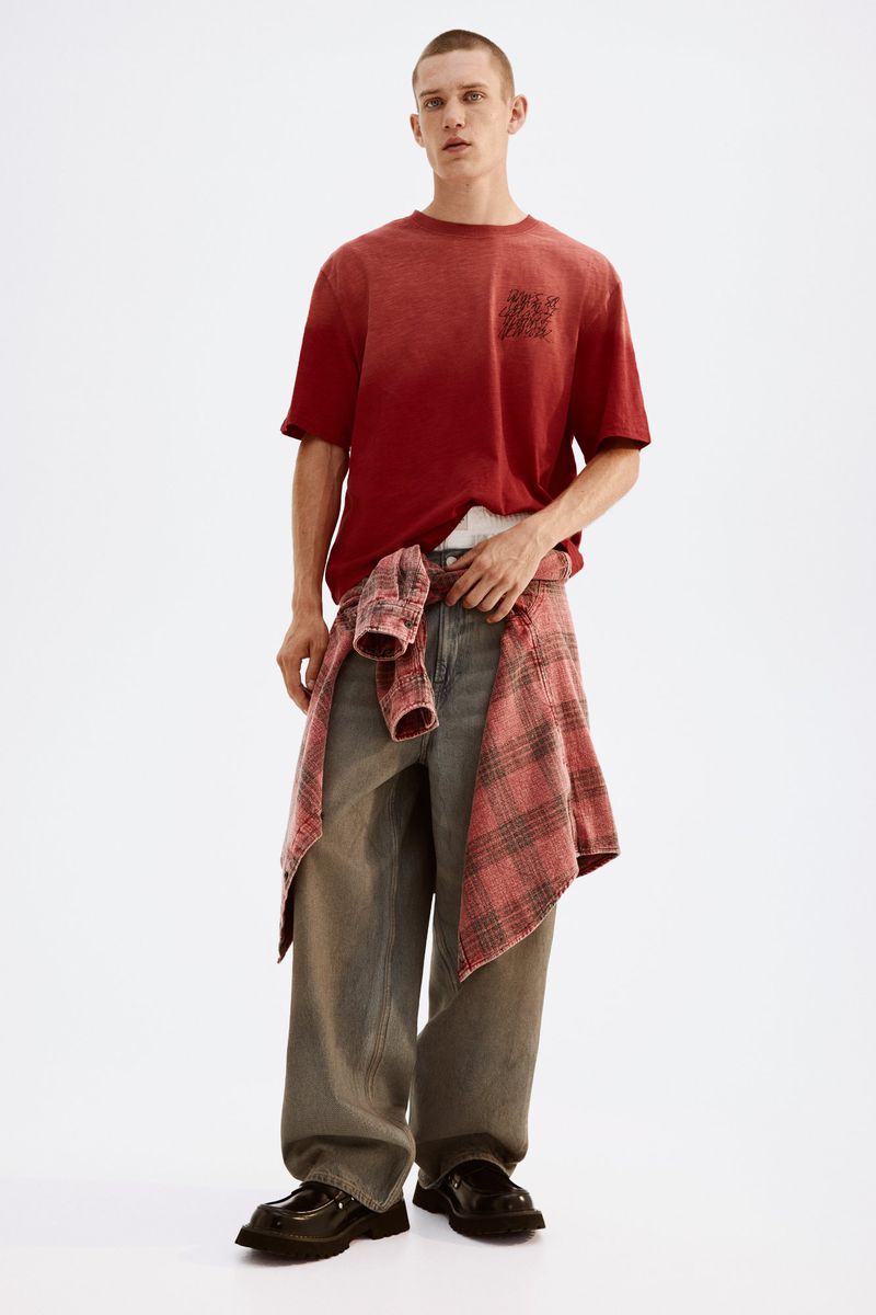 Young man in red ombre t-shirt and grey jeans, standing indoors with a plaid shirt tied around his waist, looking at the viewer.