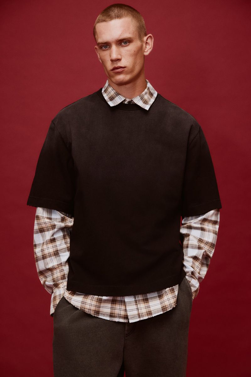 Model Jake Hodder wearing a black short-sleeve shirt over a plaid collared shirt against a deep red studio backdrop.