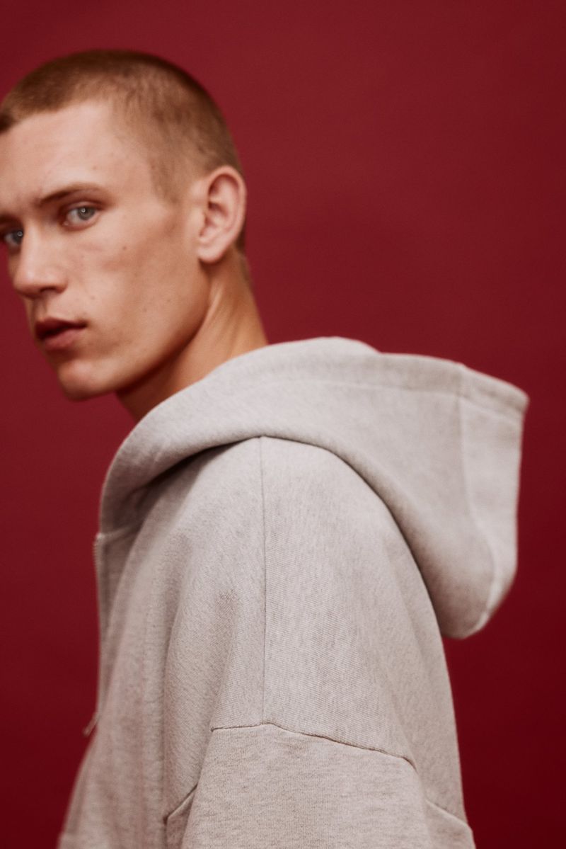Model Jake Hodder wearing an oversized light grey hooded sweatshirt against a solid deep red studio background.