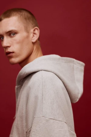 Model Jake Hodder wearing an oversized light grey hooded sweatshirt against a solid deep red studio background.