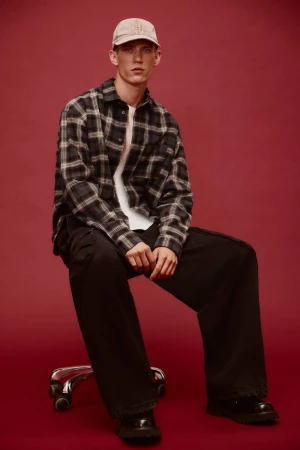 Man in plaid shirt and baseball cap sitting on a wheeled stool against a dark red background in a fashion studio setting.