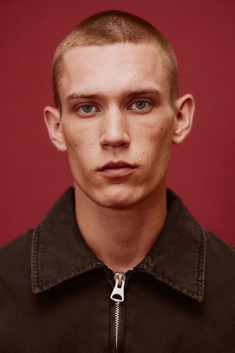 Close-up portrait of a young adult male with short hair and blue eyes wearing a dark collared shirt against a solid red background.