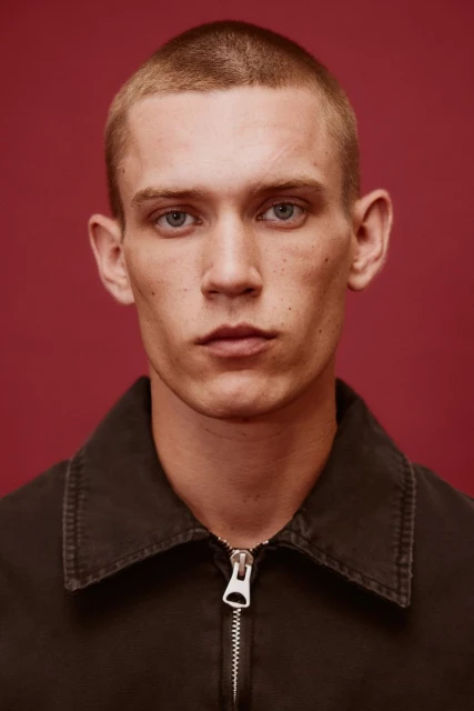 Close-up portrait of a young adult male with short hair and blue eyes wearing a dark collared shirt against a solid red background.
