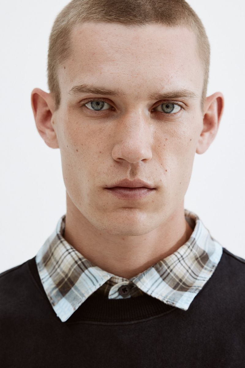 Close-up portrait of an adult male with a buzzcut, wearing a black sweater with a collared plaid shirt underneath, against a white backdrop.