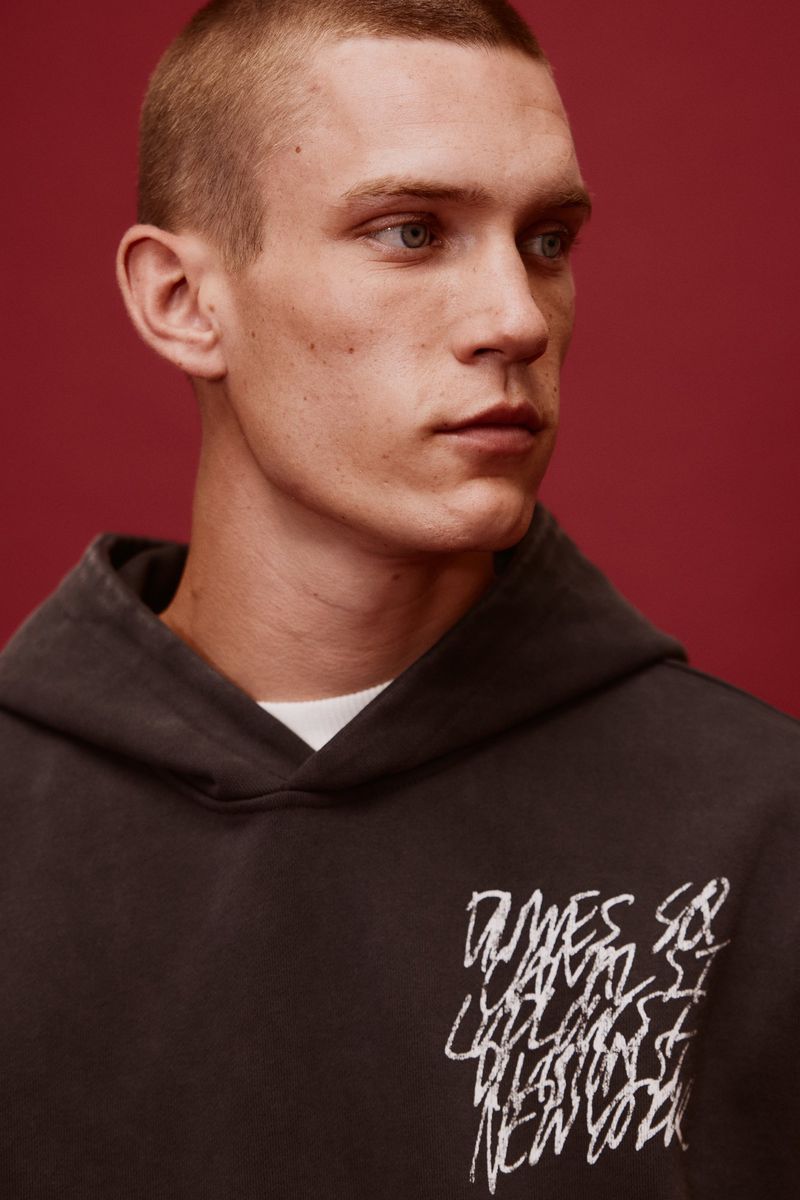 Close-up portrait of a young adult male with short hair wearing a hoodie with white graphic text against a maroon backdrop.