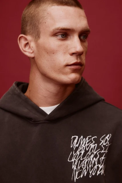 Close-up portrait of a young adult male with short hair wearing a hoodie with white graphic text against a maroon backdrop.