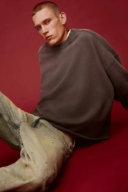 Portrait of a young adult male wearing a gray crewneck sweatshirt and light wash jeans against a dark red background.