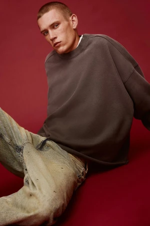 Portrait of a young adult male wearing a gray crewneck sweatshirt and light wash jeans against a dark red background.