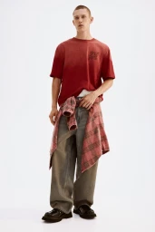 Model Jake Hodder wearing a red graphic t-shirt, plaid shirt tied at waist, and baggy grey jeans against a white backdrop.