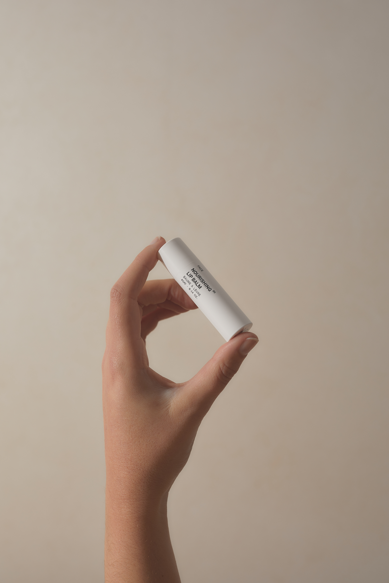 Adult human hand holds a white lip balm tube against a tan background in a simple product shot for beauty and healthcare uses.