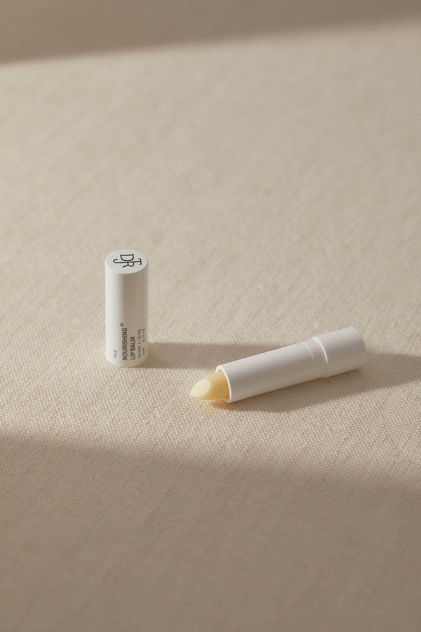 Two white tubes of lip balm sit on a textured beige surface, one open revealing the balm, in a well-lit, simple product shot.