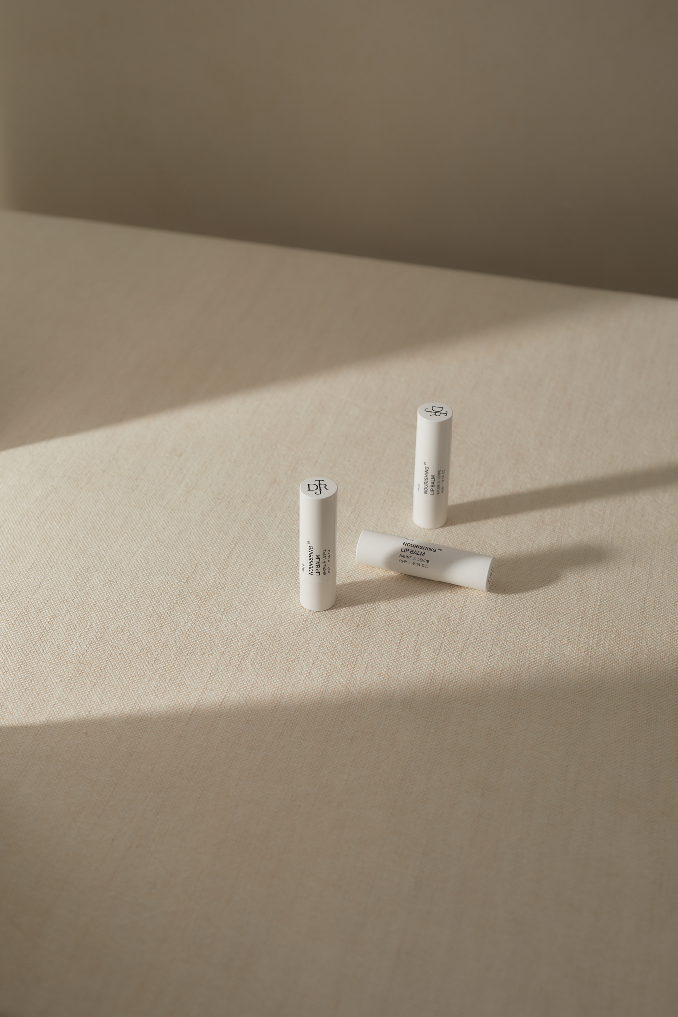 Three white lip balm tubes are positioned on a cream-colored surface, two standing upright and one lying horizontally in sunlight.