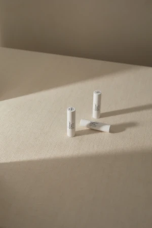 Three white lip balm tubes are positioned on a cream-colored surface, two standing upright and one lying horizontally in sunlight.