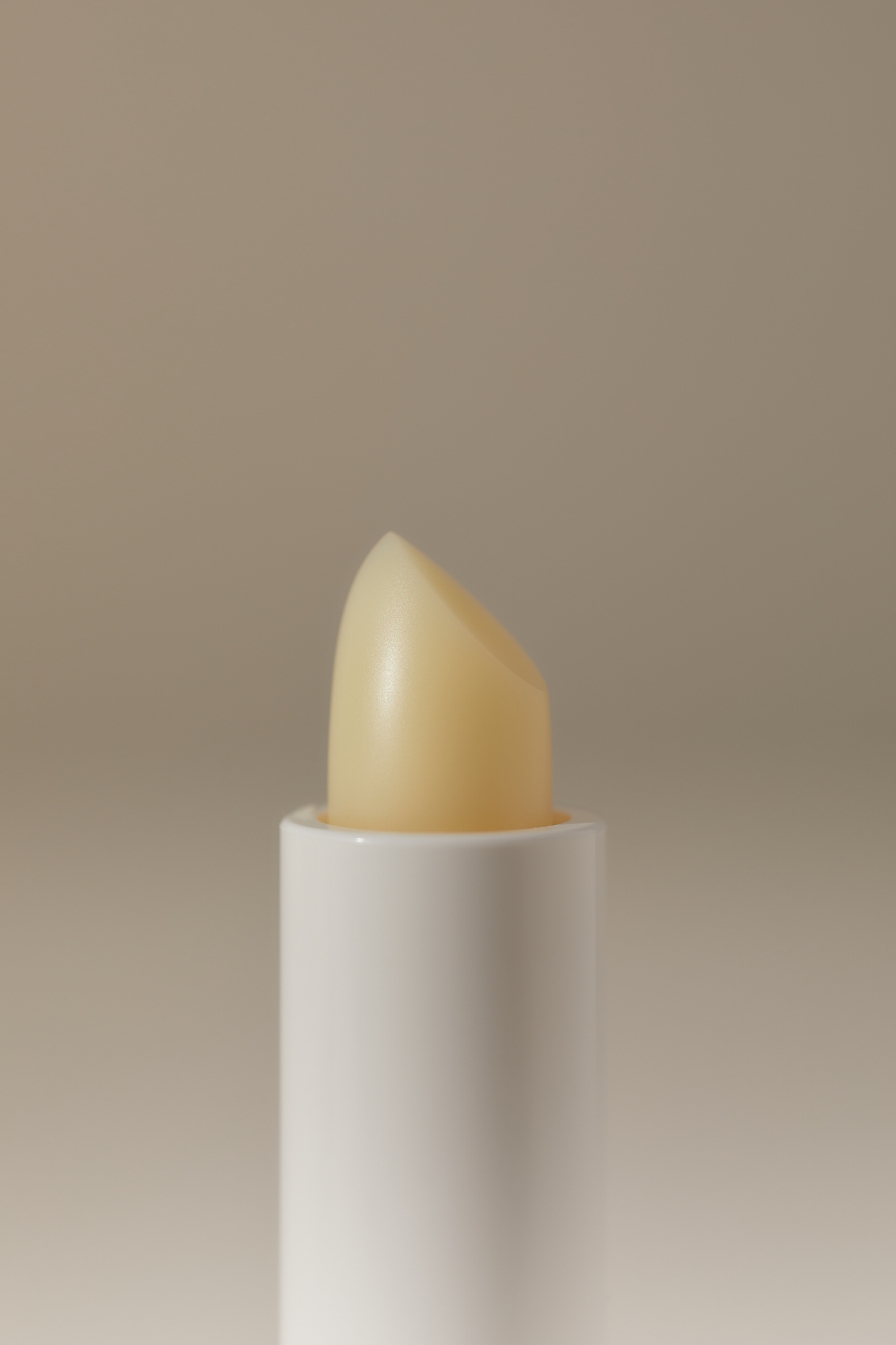 Close-up of a white tube of lip balm with the pale yellow product extended, set against a neutral beige background in a studio.