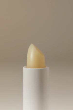 Close-up of a white tube of lip balm with the pale yellow product extended, set against a neutral beige background in a studio.