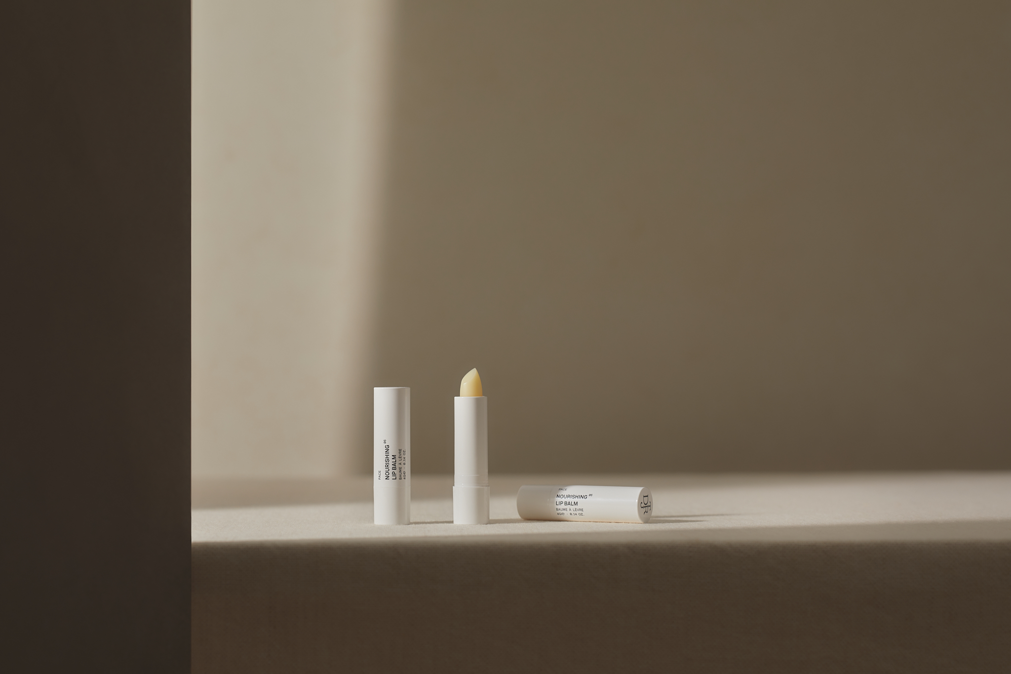 Three white lip balm tubes are arranged on a beige surface with a neutral background, one tube is open showing the balm stick.