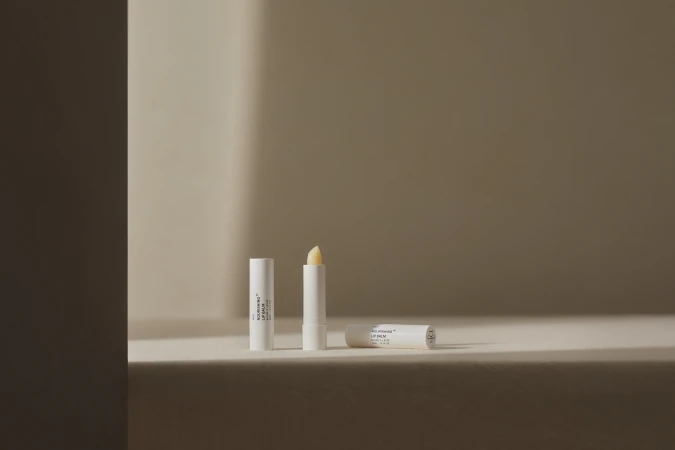 Three white lip balm tubes are arranged on a beige surface with a neutral background, one tube is open showing the balm stick.