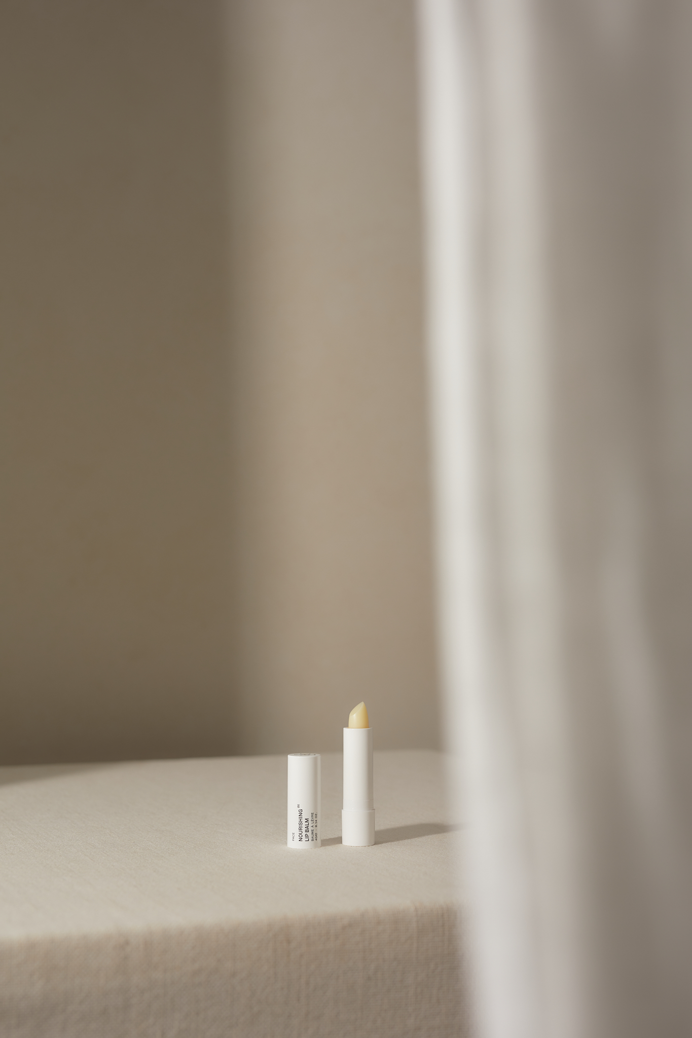 Close-up of a white lip balm tube with the cap off on a white surface, lit by natural light, with a blurred white curtain.